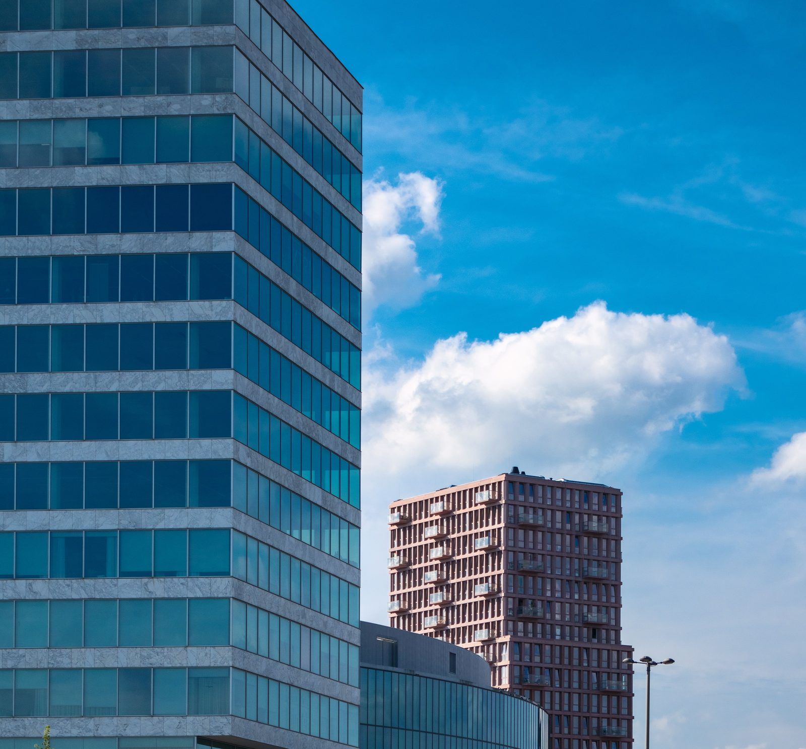 Glazen kantoortoren op de voorgrond met daarachter een roze woontoren tegen een blauwe lucht